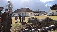 Zwei Soldaten liegen in einer kleinen Senke und haben ihre Waffen im Anschlag. Mehrere Personen beobachten die Situation.