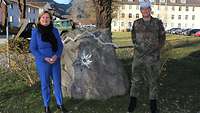 Eine Zivilistin und ein Soldat stehen vor einem Findling in einer Kaserne.