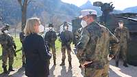 Im Vordergrund sprechen eine Zivilistin und ein Soldat miteinander, im Hintergrund stehen Soldaten und ein Fahrzeug.
