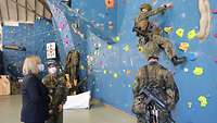 Ein Soldat steigt eine Kletterwand hoch, während eine Soldatin einer Zivilistin etwas erklärt.