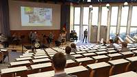 Ein großer Saal mit vereinzelten Soldaten darin. Auf einer Leinwand ist ein General am Schreibtisch zu sehen
