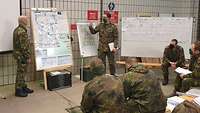 Zwei Soldaten stehen an Tafeln mit Landkarten. Davor sitzen mehrere Soldaten und machen Notizen.