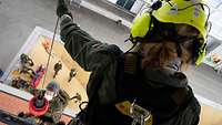 Soldatin ist in großer Höhe und hält ein Seil in der linken Hand, am Seil hängt ein Soldat mit Helm