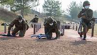 Zwei Soldaten kriechen unter flach gespannten Drähten durch. Ein dritter Soldat steht mit seiner Waffe auf und rennt weiter.