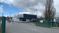 Zwei Fahrspuren, ein grünes Metalltor, dahinter ein zweistöckiges Gebäude, davor ein Wachhaus.