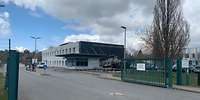 Zwei Fahrspuren, ein grünes Metalltor, dahinter ein zweistöckiges Gebäude, davor ein Wachhaus.