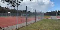 Eine rote, rechteckige Tartanfläche mit Balltor und Basketballnetz umgeben von einem hohen Zaun.