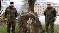 Zwei Soldaten stehen links und rechts neben einem mit Symbolen geschmückten Findling auf einer Wiese.