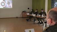 Soldaten sitzen in einem Saal verteilt an Tischen und blicken auf eine Leinwand.