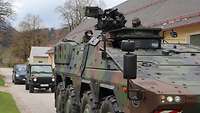 Soldaten fahren mit einem grün getarnten Radpanzer auf der Straße, es folgen Autos.