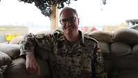 Ein Militärseelsorger in Uniform steht vor mehreren Sandsäcken und lächelt in die Kamera 