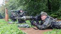 Zwei Soldaten hocken beziehungsweise liegen am Rande eines Waldes und halten eine Panzerfaust im Anschlag.
