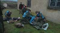 Soldaten mit blauen Schutzwesten versorgen Verletzte auf einer Wiese neben einem Gebäude
