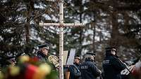 Musiker in Polizeiuniform spielen Musik auf einer Friedhofswiese, im Vordergrund ein christliches Kreuz aus Birkenholz.