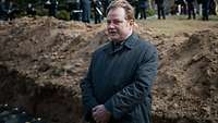 Ein Zivilist in grauem Mantel spricht auf einem Friedhof.