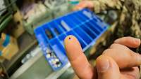 Eine sehr kleine Schraube auf einem Finger, im Hintergrund eine Kiste mit verschiedenen Schrauben 