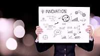 Eine Zeichnung mit der Aufschrift Innovation wird von einer Person hochgehalten.