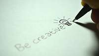 Jemand schreibt das Word Be Creativ auf ein Blatt Papier und zeichnet ein Glühlampe dahinter.
