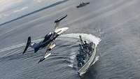 A small jet airplane flies over a grey warship at sea.