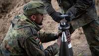 Zwei Soldaten halten mittig zwischen sich eine Granate. An der Spitze wird der Zünder eingestellt.