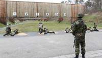 Mehrere Soldaten liegen auf Schießbahnen nebeneinander und schießen eine Übung. Hinter Ihnen steht der Leitende und beobachtet.
