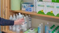 Zu sehen sind ein gut gefüllter Schrank mit Hygieneartikeln (FFP2-Masken, Desinfektionsmittel etc.) und eine greifende Hand.