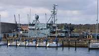 Ein Marineschiff liegt an einer Pier, im Vordergrund kleine, weiße Segelboote.