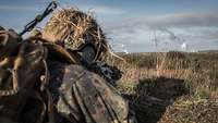 Soldaten liegen im hohen Gras in Stellung. Weiter entfernt gehen Geschosse der Artillerie nieder.