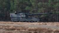 Ein Panzer in schneller Fahrt von links nach rechts vor einer Waldkante.