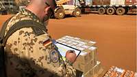 Ein Soldat steht mit einem Ordner in der Hand vor einem Anhänger und gleicht die Lieferung mit der bestellten Ware ab