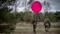 Zwei Soldaten laufen durchs Gelände. Einer hält einen roten Wetterballon in der Hand.