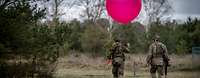 Zwei Soldaten laufen durchs Gelände. Einer hält einen roten Wetterballon in der Hand.