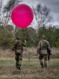Zwei Soldaten laufen durchs Gelände. Einer hält einen roten Wetterballon in der Hand.