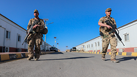 Zwei Soldaten mit G36 laufen eine Straße entlang in Richtung der Kamera. Links und rechts stehen lange Containergebäude