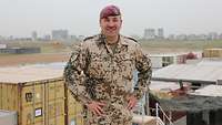 Ein Soldat hat beide Hände in die Hüfte gestemmt und lächelt in die Kamera. Im Hintergrund liegt Erbil 