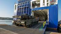 Ein Kampfpanzer in Tarnfarben rollt über eine große Rampe von einem großen blau-weißen Transportschiff.