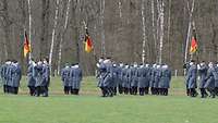 Soldaten stehen angetreten in einer Formation. Weitere Soldaten marschieren mit Fahnen zum Appell ein.