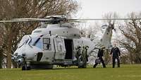 Mehrere Soldaten stehen auf einem grünen Rasen neben einem hellgrauen Hubschrauber; der Rotor dreht sich noch.