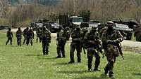 Mehrere Soldaten gehen auf einer Wiese. Im Hintergrund sind Berge und Militärfahrzeuge zu sehen.