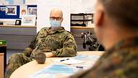 Ein General mit Mund-Nasen-Schutz sitzt an einem Tisch und unterhält sich mit einem Soldaten, der ihm gegenübersitzt