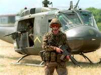 Ein Soldat in grüner Uniform steht mit dem Gewehr vor dem Hubschrauber auf einer Wiese.