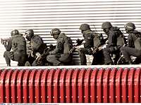 Sechs Soldaten in schwarzem Gefechtsanzug hocken hintereinander an einer silbernen Häuserwand.