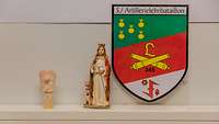 Ein Keramikengel und eine Keramikfrau stehen auf einem Regal, an der Wand ein Wappen der 5./ Artillerielehrbataillon 345 