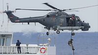Sea-Lynx-Pilot Fast Roping Ein grauer Hubschrauber schwebt über einem Schiff. Eine Person in Uniform seilt sich aus dem Hubschrauber ab