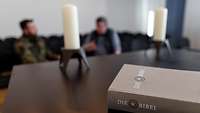 Tisch mit Bibel und Kerze, im Hintergrund ein Soldat und ein Zivilist.
