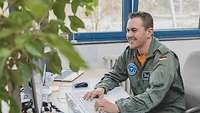Ein Mann in einem grauen Overall sitzt an einem Schreibtisch vor einem Computer