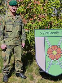 Zwei Soldaten in Uniform stehen draußen neben einem Wappen auf der grünen Wiese. 