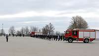 Mehrere Personen in Feuerwehruniform bei einem förmliche Appell. Daneben zwei Feuerwehrfahrzeuge.