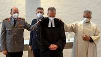 Ein Pfarrer im schwarzen Talar blickt zu Boden. Ein General, ein Polizist, ein Pfarrer legen ihm eine Hand auf die Schulter.