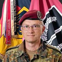 Ein Soldat mit Brille schaut in die Kamera, hinter ihm die Truppenfahne.